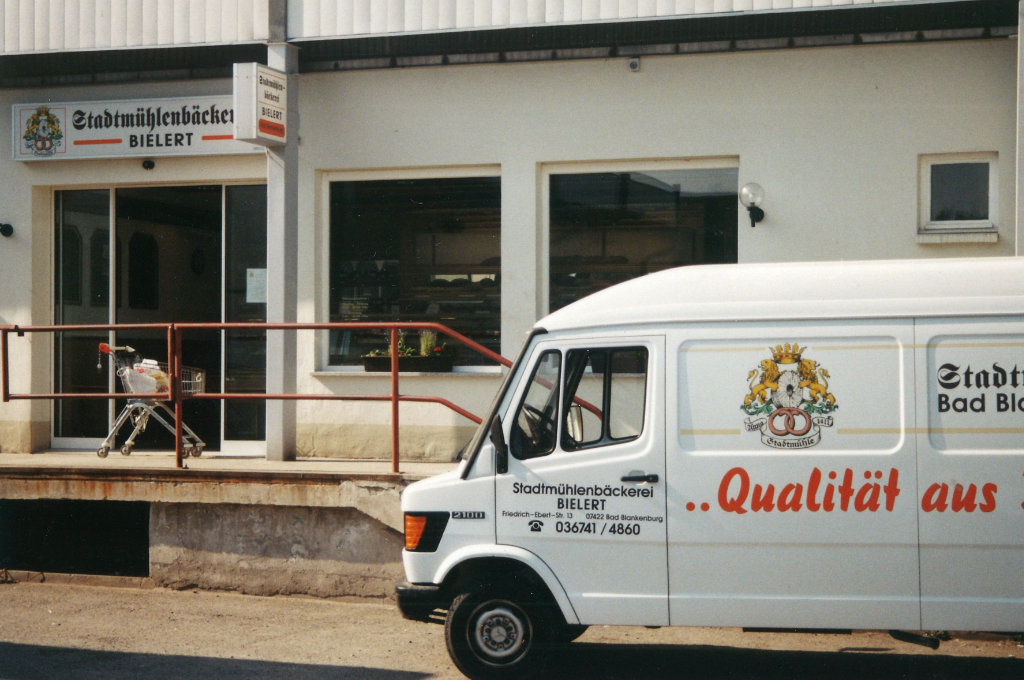 Bielert-Filiale mit Lieferfahrzeug – Qualität sichtbar gemacht Filiale der Stadtmühlenbäckerei Bielert mit weißem Lieferwagen im Vordergrund, auf dem das Wappen und die Aufschrift „Qualität aus...“ zu sehen ist.