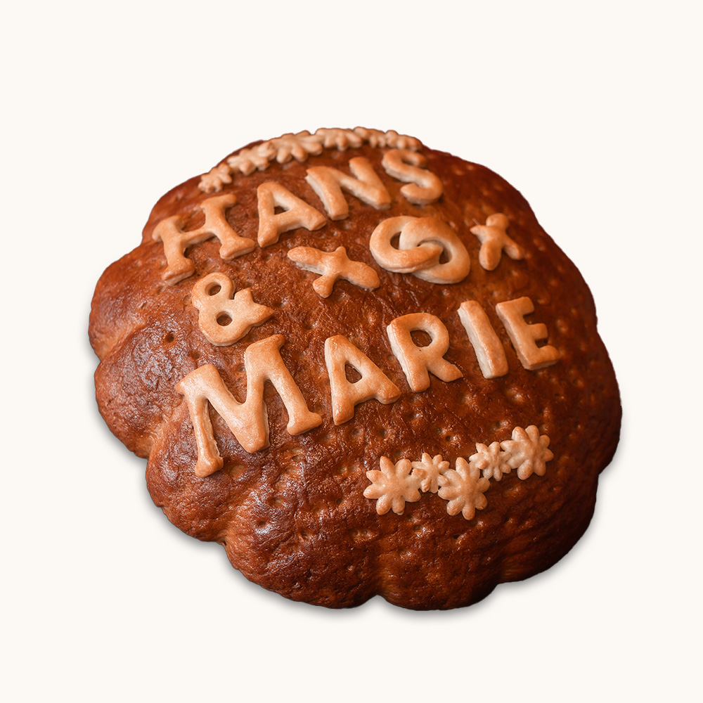 Rundes, braun gebackenes Schaubrot mit dem Namen „Hans & Marie“ und dekorativen Blümchen aus Teig.